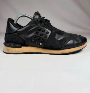 Svarta Valentino rockrunners  - Snygga svarta sneakers från Valentino med coola pyramidformade nitar på sidorna och hälen. Skorna är gjorda i en mix av läder och mocka, har svarta skosnören och en beige sula som ger en schysst kontrast. Perfekta för dig som vill sticka ut med din stil.
