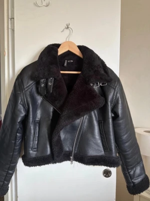 Svart biker skinnjacka med fuskpäls - Snygg svart biker skinnjacka från H&M med mjukt fuskpälsfoder och detaljerade spännen vid kragen. Jackan har sneddragkedja och två fickor.