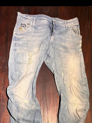 Ljusblå slitna jeans från G-Star - Säljer ett par ljusblå jeans från G-Star med slitningar och distressed detaljer på benen. Jeansen har klassisk femficksmodell, raka ben och är gjorda i bomull. Perfekta för en avslappnad och trendig look. Size 33/36