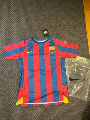 Barcelona Ronaldinho matchtröja Nike - Säljer en klassisk Barcelona matchtröja från Nike med Ronaldinho och nummer 10 på ryggen. Tröjan har blå och röda vertikala ränder, klubbmärke på bröstet och gula detaljer. Perfekt för dig som älskar fotboll och vill hylla en legend.