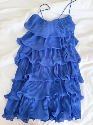 Blå volangklänning  - Supersöt blå kortklänning med tunna axelband och flera lager volanger. Klänningen är plisserad och har en luftig, flowy känsla som passar perfekt till sommaren. 
