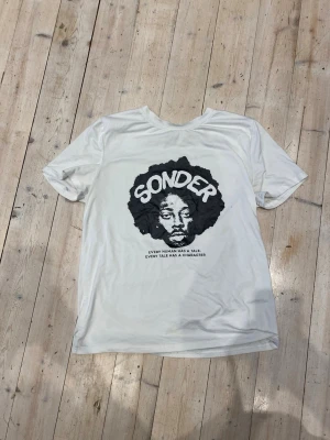 Vit t-shirt med svart tryck SONDER - Vit t-shirt med stort svart tryck på bröstet där det står 'SONDER' och texten 'Every human has a tale. Every tale has a character.' Rund halsringning och normal passform. Perfekt för dig som gillar grafiska prints.