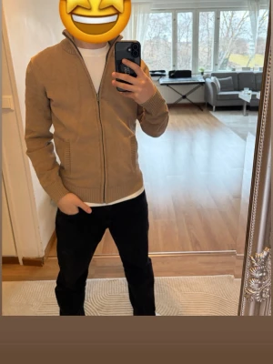 Beige ziptröja - Säljer en beige stickad ziptröja med ribbade muddar och krage. Tröjan har två fickor framtill och dragkedja hela vägen. Perfekt för lager på lager och enkel att matcha med olika outfits.