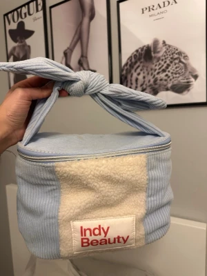 Indy Beauty ljusblå necessär - Supersöt necessär från Indy Beauty i ljusblått manchestertyg med vita teddy-detaljer. Har en knytbar handtag upptill och dragkedja runt om. Perfekt för smink eller småprylar. Märkets logga i rött framtill. Blir ren i tvätten 