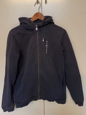 Mörkblå ziphoddie  från Sail Racing - Snygg mörkblå hoodie från Sail Racing med dragkedja framtill och en bröstficka med logga. Hoodien har justerbar huva och två sidofickor. Tillverkad i mjukt material som känns skönt mot huden. Perfekt för en avslappnad och sportig look.
