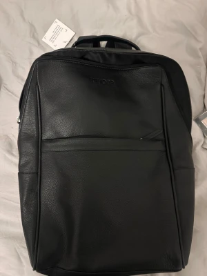 Svart ryggsäck i konstläder från Kayoba - Stilren svart ryggsäck från Kayoba i konstläder med minimalistisk design. Ryggsäcken har justerbara axelband, bärhandtag och en stor ytterficka framtill. Perfekt för laptop och vardagsprylar. Snygg och enkel look som passar till det mesta.