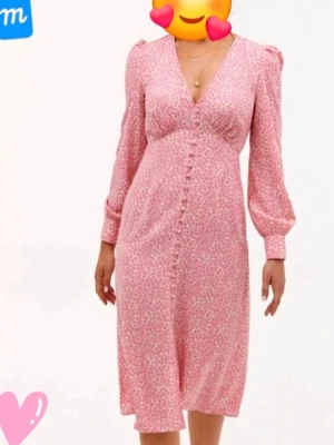 Rosa blommig klänning med knappar - Supersöt rosa klänning med små blommor, v-ringning och långa puffärmar. Klänningen har knappar framtill och markerad midja för en feminin siluett. Perfekt för vår och sommar, och ger en romantisk vibe.