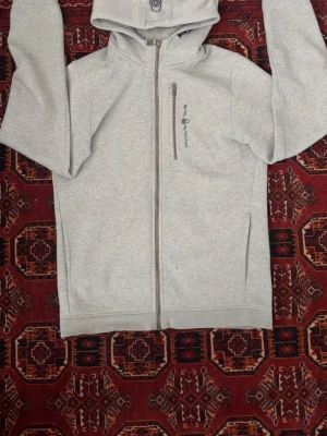 Ljusgrå hoodie från Sail Racing - Ljusgrå hoodie från Sail Racing med dragkedja framtill och en bröstficka med vertikal dragkedja. Mjuk insida i fleece och klassisk huva med knappdetalj. Perfekt för chill dagar eller när du vill ha något bekvämt och stilrent.