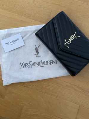 Svart väska från Yves Saint Laurent - Snygg svart kuvertväska från Yves Saint Laurent i quiltat läder med guldigt YSL-emblem på framsidan. Klassisk och stilren design med kuvertformad lock och elegant struktur. Perfekt accessoar för att lyfta vilken outfit som helst.