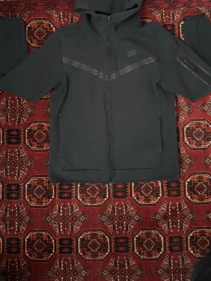 Svart Nike hoodie med dragkedja - Säljer en svart hoodie från Nike med dragkedja framtill och huva. Hoodien har två stora fickor, diskreta reflexdetaljer över bröstet och en extra ficka på ena ärmen. Tillverkad i mjukt material som känns skönt mot huden. Perfekt för en sportig och avslappnad stil.