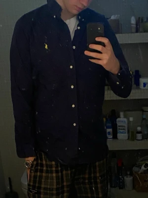 Mörkblå skjorta från Ralph Lauren - Klassisk mörkblå skjorta från Ralph Lauren med button-down krage och vita knappar. Skjortan har lång ärm och den ikoniska gula logotypen broderad på bröstet. Tillverkad i bomull och passar perfekt till en avslappnad men ändå stilren look.