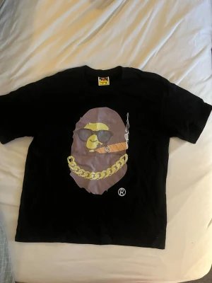 Svart Bape t-shirt med tryck - Cool svart t-shirt från Bape med stort tryck av en apa med solglasögon, guldkedja och cigarr på bröstet. Perfekt statement-plagg för dig som gillar streetwear och unika motiv.