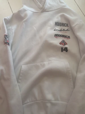 Vit hoodie från Hoodrich med tryck - Snygg vit hoodie från Hoodrich med flera tryck och broderade detaljer i svart och rött. Stor Hoodrich-logga på huvan, patch på ärmen och coola grafiktryck på bröstet. Klassisk känguruficka framtill och mjukt material som känns skönt mot huden.