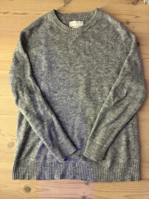 Grå stickad tröja från Samsøe - Mysig grå stickad tröja från Samsøe  med rund halsringning och ribbade muddar vid ärmslut och nederkant. Tröjan har en klassisk rak passform och är tillverkad i ett mjukt, melerat material som passar perfekt till kyliga dagar.