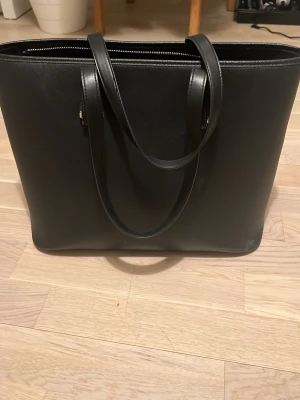 Svart axelväska i skinn - Stilren svart axelväska i slätt skinn med två långa handtag och dragkedja upptill. Väskan har en klassisk, fyrkantig form och passar perfekt för dig som vill ha plats för både laptop och vardagsprylar. Enkel och tidlös design som funkar till allt.