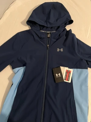 Blå hoodie från Under Armour - Snygg blå hoodie från Under Armour med dragkedja och huva. Hoodien har en mörkblå bas med ljusblå paneler på sidorna och det klassiska Under Armour-loggan på bröstet. Tillverkad i ett mjukt och stretchigt material som passar perfekt till en aktiv vardag.