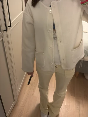 Vit cardigan jacka från RYVLS - Stilren vit cardigan jacka från RYVLS med rund halsringning, stora framfickor och knäppning med knappar. Jackan har en mjuk känsla och enkel design som passar perfekt till många outfits.