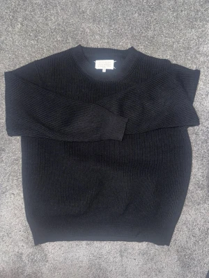 Svart stickad tröja Maison Margiela - Säljer en svart stickad tröja från Maison Margiela. Tröjan har rund halsringning, ribbstickade muddar och är gjord i ett tjockare material som känns riktigt lyxigt. Perfekt för dig som gillar minimalistisk och stilren design.