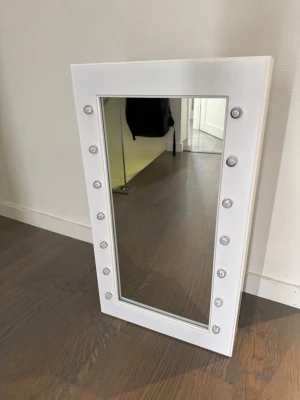 Vit sminkspegel med LED-belysning - Snygg rektangulär sminkspegel med vit ram och inbyggda LED-lampor runt spegelglaset. Perfekt för att fixa sminket med jämnt ljus. Spegeln har en modern och stilren design som passar i alla rum. Tillverkad i plast och glas.