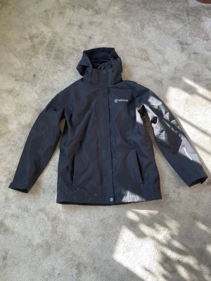 Svart skal/regnjacka från Tenson - Snygg svart skal/regnjacka från Tenson med huva och dragkedja framtill. Jackan har två sidofickor och justerbar huva. Materialet är slitstarkt och vattenavvisande, perfekt för utomhusaktiviteter och sport. Tenson-logga på bröstet ger en sportig look.