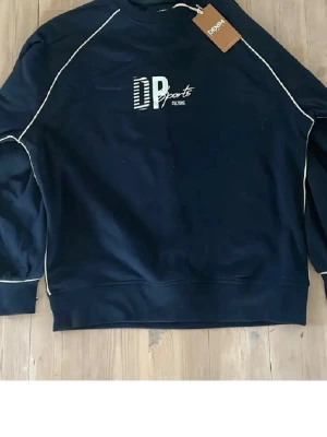 Mörkblå sweatshirt DP Sports - Mörkblå sweatshirt från DP Sports med vita kontrastsömmar och tryckt logga på bröstet. Tröjan har rund halsringning, långa ärmar och ribbade muddar vid ärmslut och nederkant. Perfekt för en sportig och avslappnad stil.