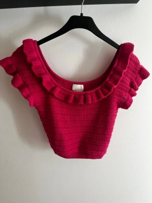 Rosa stickad croppad topp H&M - Supersöt rosa stickad croppad topp från H&M med volangdetaljer runt halsringningen och axlarna. Toppen är ärmlös och har en tight passform som framhäver figuren. Perfekt för dig som vill ha en färgglad och trendig look.