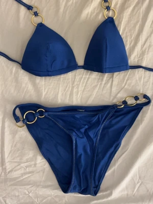 Blå bikini med guldringar - Snygg blå bikini med triangeltopp och matchande trosa. Båda delarna har dekorativa guldfärgade ringar som detaljer. Bikinin har smala band och en enkel, stilren design som känns modern och fräsch. Storlek är oklart men passar mig ändå sådär som är S men är lite förstor men går att knyta. Använd fåtal gånger.