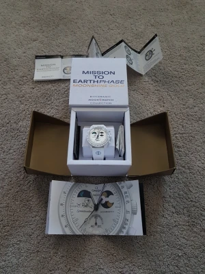 Omega x Swatch MoonSwatch vit klocka - Säljer en Omega x Swatch MoonSwatch Mission to Earthphase Moonshine Gold Snoopy. Klockan är vit med biokeramiskt material, har månfaskomplication och detaljer i guld. Snygg och futuristisk design med vit rem och tydliga svarta markeringar på urtavlan.