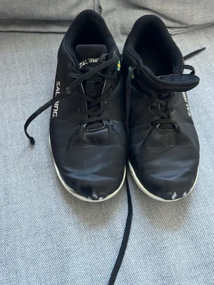 Svarta Salming inomhusskor - Säljer ett par svarta inomhusskor från Salming med vita detaljer och snörning. Skorna har låg profil, vit sula och är tillverkade i syntetmaterial. Perfekta för innebandy eller andra inomhussporter.