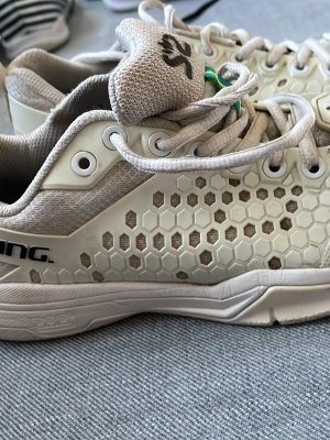 Vita Salming sneakers med hexamönster - Snygga vita sneakers från Salming med hexagonmönstrad mesh och beige detaljer. Skorna har vita snören, låg profil och en liten svensk flagga på plösen. Perfekta för dig som gillar sportig och clean stil.