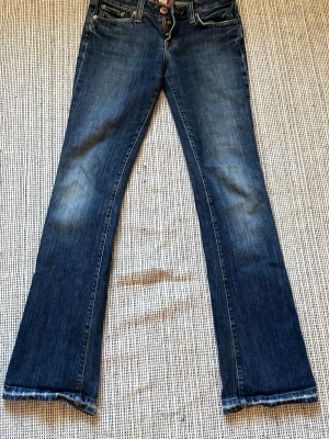 Blå bootcut jeans från Lucky Brand - Säljer ett par klassiska blå bootcut jeans från Lucky Brand med snygga slitningar och kontrastsömmar. Jeansen har dragkedjegylf, fem fickor och är tillverkade i denim med en skön känsla. Perfekta för en avslappnad och trendig look.