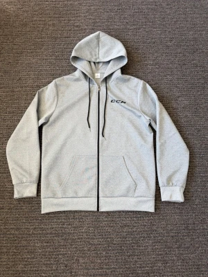 Ccm hoodie - Snygg grå zip hoodie från CCM med svart logga på bröstet. Hoodien har dragkedja framtill, två fickor och justerbar huva med svarta snören. Tillverkad i mjukt sweatshirtmaterial som är skönt att ha på sig. Perfekt för chill dagar eller till träningen.