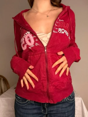 Brugundyfärgad hoodie med tryck och dragkedja❤️ - Snygg röd hoodie från U.S. Sports med vit text och siffran 05 tryckt på bröstet. Hoodien har dragkedja framtill, två fickor och en klassisk huva. Tillverkad i mjukt bomullsmaterial och har ribbade muddar vid ärmslut och nederkant. Perfekt för en avslappnad streetstyle.❤️