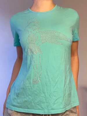 Turkos tingeling T-shirt🩵 - Snygg turkos T-shirt med korta ärmar och ett stort motiv av Tingeling framtill, gjort av glittriga stenar. T-shirten har en klassisk passform och rund halsringning. Perfekt för dig som gillar Disney och vill sticka ut lite extra.🩵