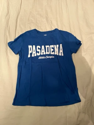 Blå Pasadena t-shirt med tryck - Snygg blå t-shirt med stort vitt tryck 'PASADENA' och Athletic Champion på bröstet. Klassisk rund hals och korta ärmar. Perfekt för en sportig och avslappnad stil. Tillverkad i mjuk bomull för skön komfort.