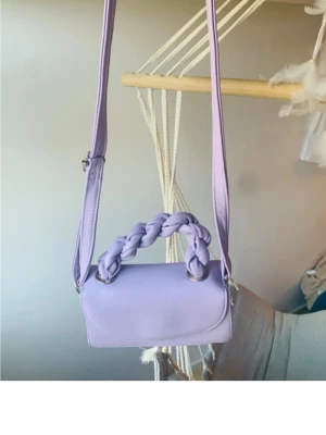 Ljuslila handväska med flätat handtag - Trendig ljuslila handväska med justerbar axelrem och ett snyggt flätat handtag. Väskan har en enkel, stilren design och är tillverkad i ett slätt material som liknar konstläder. Perfekt för dig som vill ha en färgklick till din outfit.