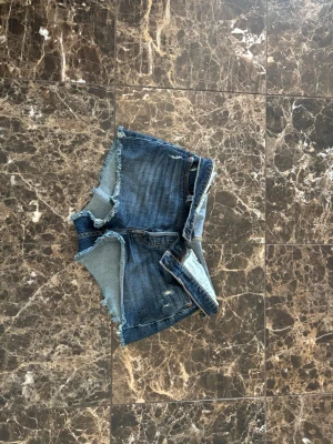 Blå Levi’s shorts - Säljer ett par blå jeansshorts med rå, fransig kant och slitna detaljer. Klassisk femficksmodell med knapp och medelhöga. Perfekta för varma dagar och ger en avslappnad look. Använd 2-3 gånger. Köpta helt nya.