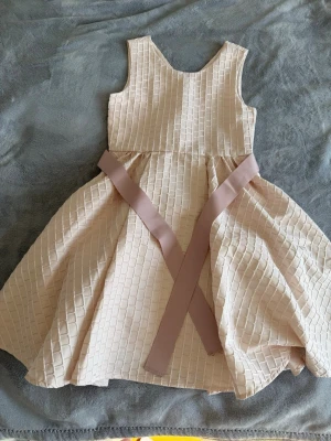 Beige festklänning med rosett - Supersöt beige klänning med strukturerat rutmönster och vid kjol. Klänningen har ärmlös design och ett brett band i midjan som kan knytas till en rosett bak. Perfekt för speciella tillfällen och ger en elegant look.