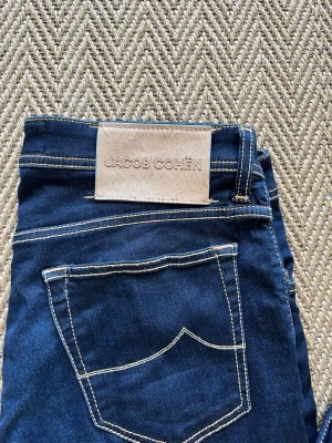 Jacob Cohën jeans - Snygga mörkblå jeans från Jacob Cohën med kontrastsömmar och klassisk femficksmodell. Jeansen har en beige patch med logga bak i midjan och är tillverkade i ett mjukt denimtyg som ger en skön passform. Perfekta för en trendig och avslappnad stil. Stolek 32 nick slim