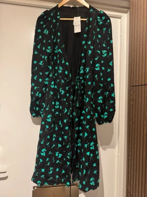 Svart blommig omlottklänning - Svart kortklänning med grönt blommönster och omlottmodell. Klänningen har långa ärmar med lätt puff och knytband i midjan för en snygg passform. Perfekt för dig som gillar en feminin och trendig stil.