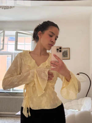 Gul blommig omlottblus med volang - Säljer en ljusgul omlottblus med subtilt blommönster och stora volangdetaljer vid halsen och ärmsluten. Blusen har långa, vida ärmar och knyts i sidan för en snygg passform. Perfekt för dig som gillar romantiska och trendiga plagg.