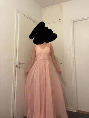 Ljusrosa långklänning med spets - Elegant ljusrosa långklänning med spetsdetaljer upptill och v-ringad hals. Kjolen är i ett flytande, tunt material som ger ett romantiskt intryck. Perfekt för bal eller festliga tillfällen. Klänningen har ärmlös design och markerad midja.
