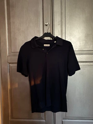 Svart piké från Jack & Jones - Klassisk svart pikétröja från Jack & Jones med krage och knappar framtill. T-shirten har korta ärmar och är tillverkad i mjuk bomull, perfekt för en clean och enkel stil.