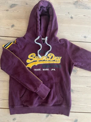 Vinröd hoodie från Superdry - Snygg vinröd hoodie från Superdry med gul och vit logga på bröstet. Klassisk ficka framtill och justerbar huva med vita snören. Mjuk insida och ribbade muddar. Passar perfekt inför våren!!🙌🙌