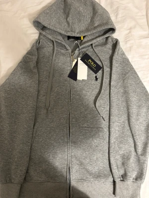 Grå zip hoodie från Polo Ralph Lauren - Snygg gråmelerad hoodie från Polo Ralph Lauren med dragkedja framtill, huva med dragsko och ribbade muddar. Klassisk liten logga broderad på bröstet. Perfekt för en chill och stilren look.