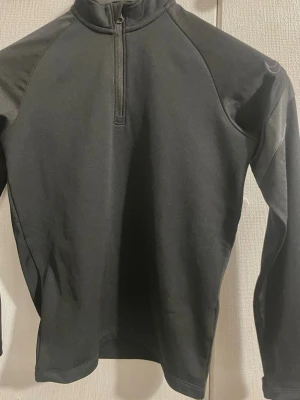 Koszulka treningowa Nike/ Täningströja Nike: junior - Svart långärmad träningströja från Nike med halv dragkedja framtill och hög krage. Tillverkad i ett stretchigt funktionsmaterial som passar perfekt för löpning eller gym. Diskret Nike-logga på ryggen och reflekterande detalj på ärmen. Storleken på denna tröja är S för juniorer.