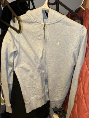 Ljusblå zip hoodie från Polo Ralph Lauren - Ljusblå hoodie med dragkedja från Polo Ralph Lauren. Klassisk design med huva, fickor framtill och den ikoniska broderade loggan på bröstet. Tillverkad i mjuk bomullsblandning som är skön att ha på sig. Perfekt för en avslappnad och stilren look. Passar S- M