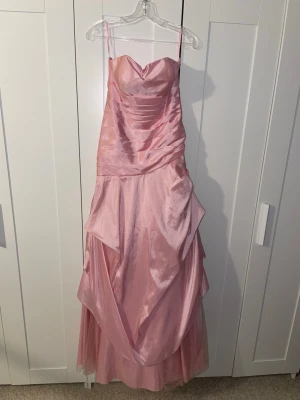 Rosa balklänning med snörning bak - Snygg rosa axelbandslös balklänning med draperad kjol. Klänningen har vacker snörning i ryggen för förbättrad passform och ett feminint fall. Perfekt för bal eller festliga tillfällen.