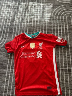 Liverpool matchtröja Nike Salah 11 - Röd Liverpool matchtröja från Nike med tryck av M. Salah och nummer 11 på ryggen. Tröjan har vita och turkosa detaljer, klubbmärke och sponsorloggor på bröstet samt Premier League-märke på ärmen. Tillverkad i lätt och ventilerande material.