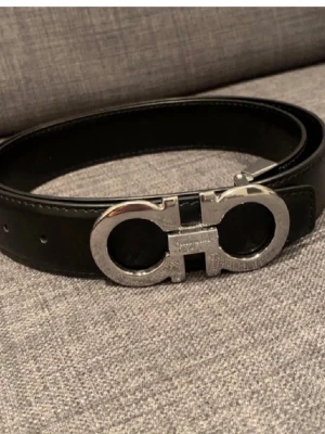  Salvatore Ferragamo - Snyggt svart skärp från Salvatore Ferragamo med ikoniskt dubbel-Gancini-spänne i silverfärgad metall. Bältet är gjort i slätt skinn och har en stilren, klassisk look som passar till många outfits. Perfekt accessoar för dig som gillar exklusiva detaljer.